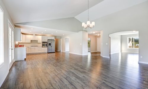 Spacious rambler home interior with vaulted ceiling over glossy laminate floor. Empty light filled dining or living space adjacent to new white kitchen room features pale grey walls. Northwest, USA